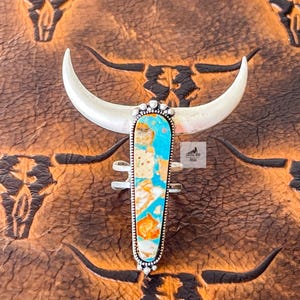 May include: A silver-toned ring with a turquoise and orange stone, set in a decorative frame. The ring is shaped like a bull's head, with curved horns. The background features a repeating pattern of bull skull silhouettes.