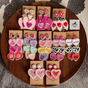 Może przedstawiać: Zestaw kolczyków z koralików w stylu walentynkowym. Kolczyki mają kształt serc, butelek perfum i serduszek z cukierków. Kolczyki są w różnych kolorach, w tym różowym, czerwonym, białym i złotym. Kolczyki są prezentowane na drewnianej powierzchni.