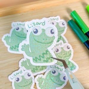 May include: A collection of green lizard stickers with the word "Lizard" above each cartoon lizard. The stickers have a glittery finish and are being handled with tweezers. Green and blue markers are visible in the background.