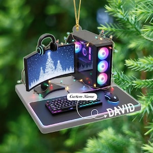 May include: A gaming computer ornament with a curved monitor displaying a snowy Christmas tree scene. The ornament includes a keyboard, mouse, and headset. The name "DAVID" is written on the mat. The ornament is decorated with colorful lights.
