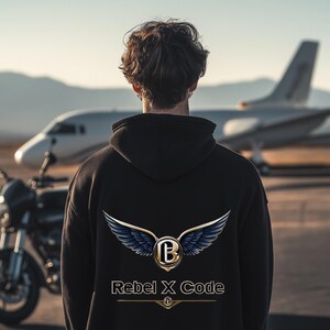 May include: A person wearing a black hoodie with a gold and blue logo that says "Rebel x Code". The logo features a stylized letter "B" with wings. The person is standing in front of a private jet.