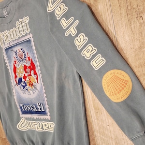 May include: A blue-grey sweatshirt with white lettering and a stamp design. The stamp features a coat of arms and the text "TONGA $1 CULTURE". The sleeve has the word "Valuable" in white and a circular patch.