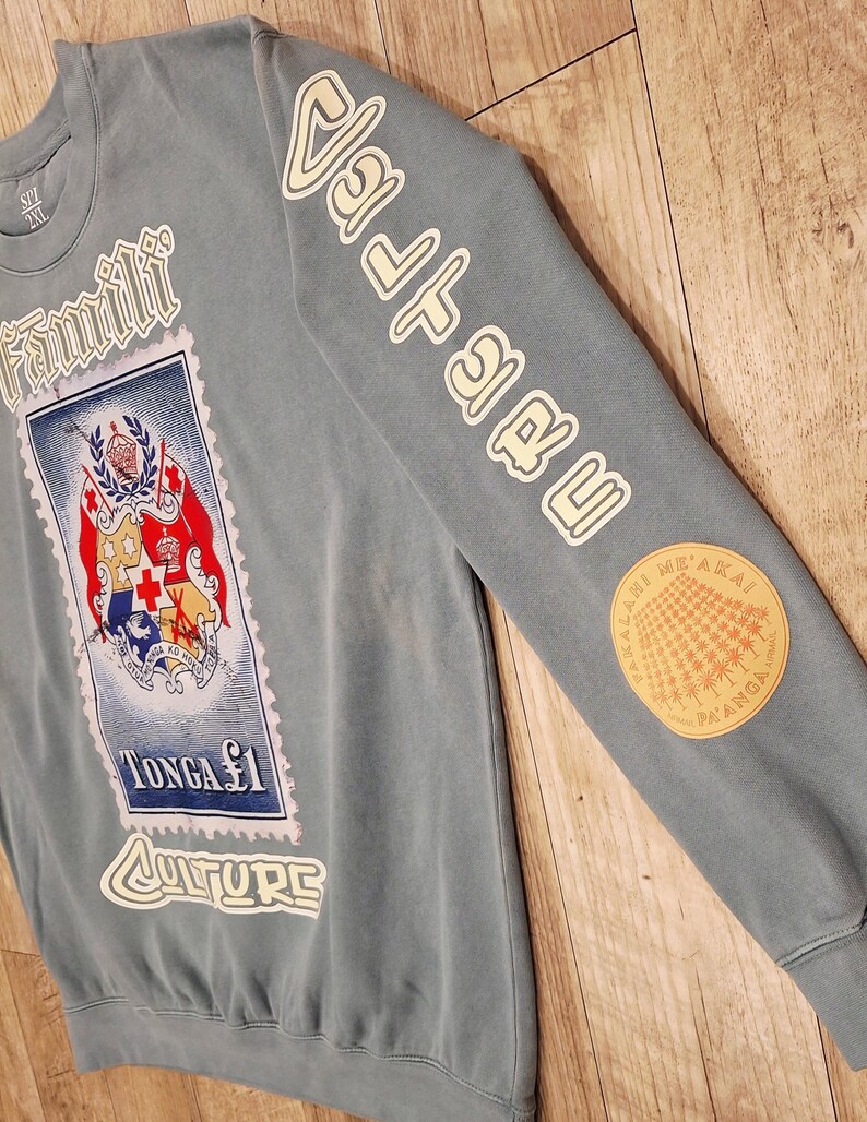 May include: A blue-grey sweatshirt with white lettering and a stamp design. The stamp features a coat of arms and the text "TONGA $1 CULTURE". The sleeve has the word "Valuable" in white and a circular patch.