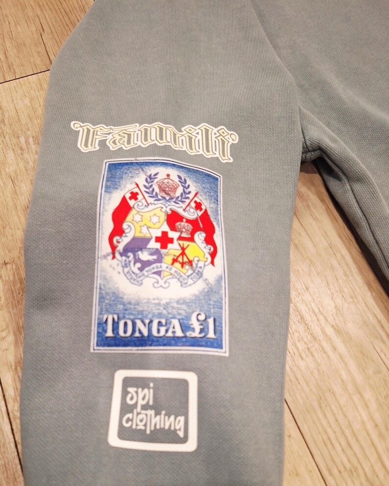 May include: A grey sweatshirt sleeve with three white patches. The top patch reads "Familii". The middle patch features a crest with red, white, and yellow elements. The bottom patch reads "spi clothing".