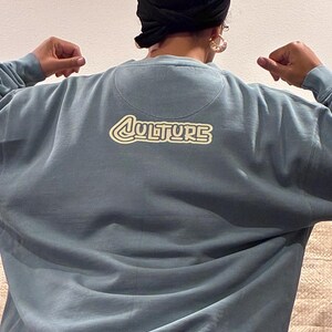 May include: A blue-grey sweatshirt with the word "CULTURE" in a retro-style font on the back. The sweatshirt has a crew neck and long sleeves. The image shows the back of the sweatshirt.