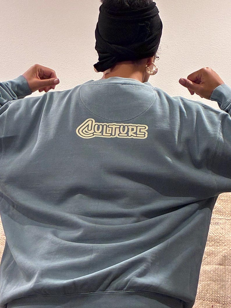 May include: A blue-grey sweatshirt with the word "CULTURE" in a retro-style font on the back. The sweatshirt has a crew neck and long sleeves. The image shows the back of the sweatshirt.