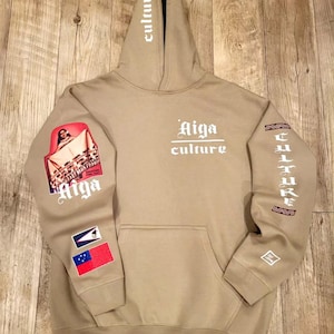 May include: A tan hoodie with the words "Aiga Culture" printed on the front. The sleeves have a graphic print of a woman in a red dress and the words "Aiga" and "Culture" printed vertically.