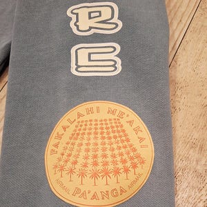 May include: A light blue sweatshirt sleeve with white outlined text that reads "URB" vertically. A circular patch features a palm tree design and the text "FAKALA HI ME'AKAI AIRMAIL PA'ANGA AIRMAIL".