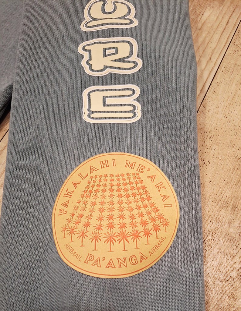 May include: A light blue sweatshirt sleeve with white outlined text that reads "URB" vertically. A circular patch features a palm tree design and the text "FAKALA HI ME'AKAI AIRMAIL PA'ANGA AIRMAIL".