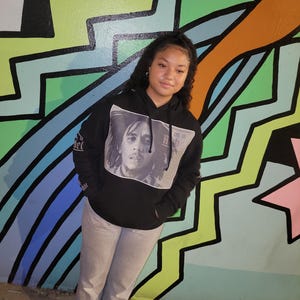 May include: A black hoodie featuring a grayscale image of Bob Marley. The hoodie has a drawstring hood and long sleeves. The person is wearing gray jeans and black and white sneakers. The background is a colorful mural with geometric shapes.
