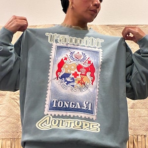 May include: A blue-green sweatshirt with a large postage stamp graphic. The stamp reads "Tonga &pound;1" and "Culture", and features a crest with red, white, and blue details. The sweatshirt has long sleeves and a crew neck.