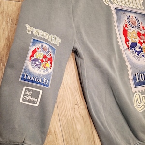 May include: A faded blue sweatshirt with the word "Famili" in white script. The sweatshirt features a large stamp-like graphic with the Tonga coat of arms and the text "TONGA &pound;1". The sleeve has a smaller version of the stamp graphic and the "spi clothing" logo.