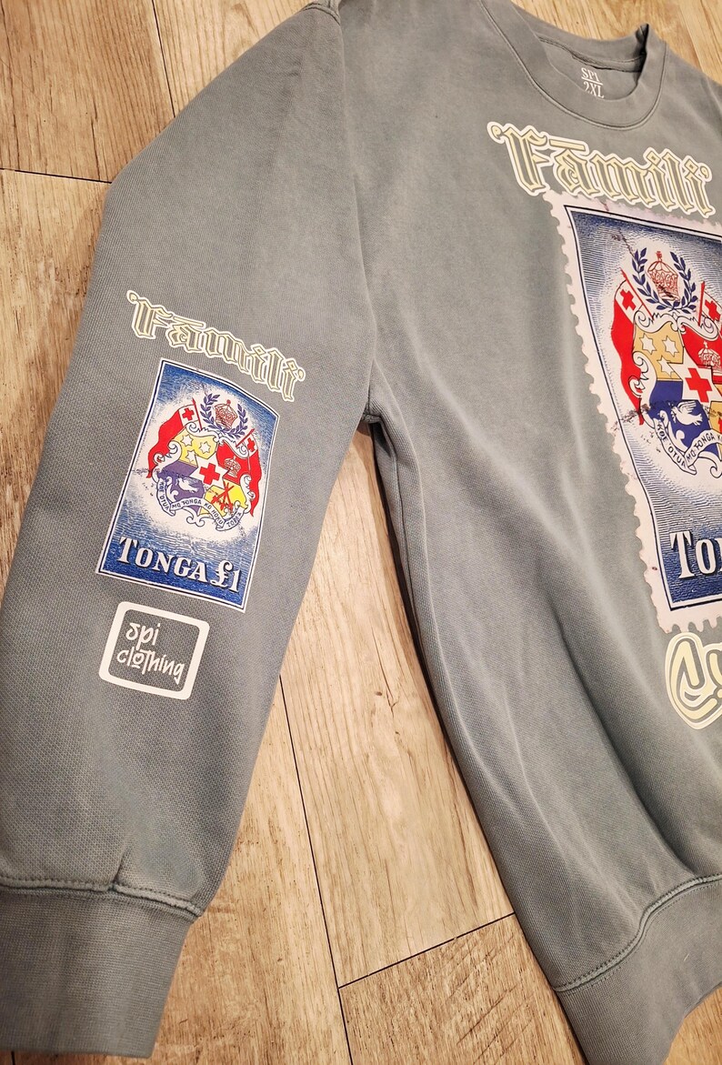 May include: A faded blue sweatshirt with the word "Famili" in white script. The sweatshirt features a large stamp-like graphic with the Tonga coat of arms and the text "TONGA &pound;1". The sleeve has a smaller version of the stamp graphic and the "spi clothing" logo.
