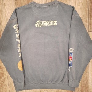 May include: A faded blue sweatshirt featuring the word "CULTURE" in a white outlined font on the back. The left sleeve displays the word "vacation" in white. The right sleeve has a cartoon graphic.