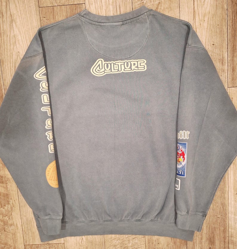 May include: A faded blue sweatshirt featuring the word "CULTURE" in a white outlined font on the back. The left sleeve displays the word "vacation" in white. The right sleeve has a cartoon graphic.