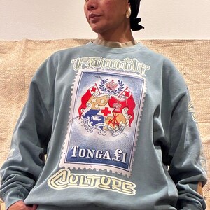 May include: A light blue sweatshirt with a large postage stamp graphic. The stamp reads "TONGA &pound;1" and "CULTURE", with a design including a crown and cross. The word "Family" is printed above the stamp in a stylized font.
