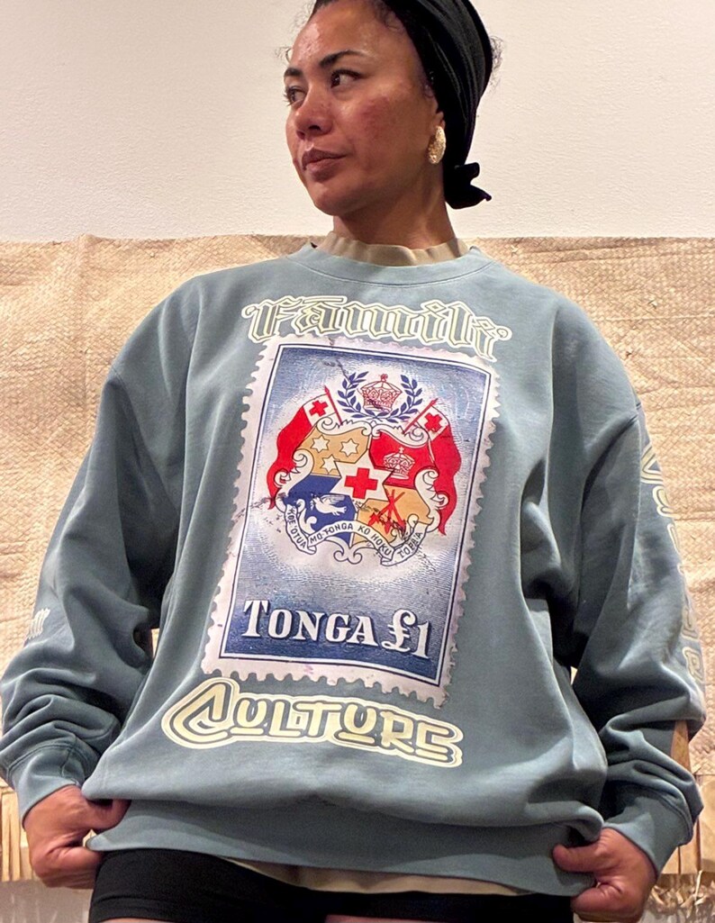 May include: A light blue sweatshirt with a large postage stamp graphic. The stamp reads "TONGA &pound;1" and "CULTURE", with a design including a crown and cross. The word "Family" is printed above the stamp in a stylized font.