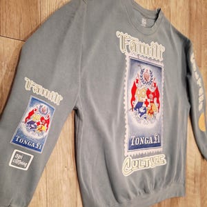 May include: A blue-grey sweatshirt with the word "Famili" in white script. The shirt features a postage stamp design with the words "TONGA &pound;1" and "CULTURE". The sleeve has a patch that says "api clothing".