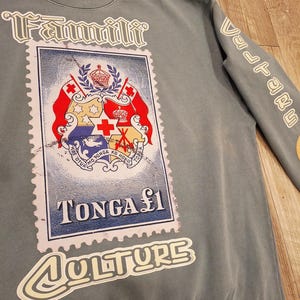 May include: A grey sweatshirt with a large graphic of a postage stamp featuring the Tonga coat of arms. The stamp reads "TONGA &pound;1". The word "Fammilli" is above the stamp, and "Culture" is below. The sleeve has the word "Vulture".