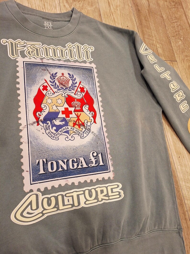 May include: A grey sweatshirt with a large graphic of a postage stamp featuring the Tonga coat of arms. The stamp reads "TONGA &pound;1". The word "Fammilli" is above the stamp, and "Culture" is below. The sleeve has the word "Vulture".