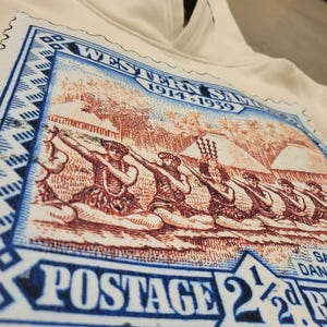 May include: A white hoodie with a vintage-style postage stamp graphic featuring a scene of people dancing. The stamp reads "Western Samoa 1914-1939" and "Postage 2 1/2d".