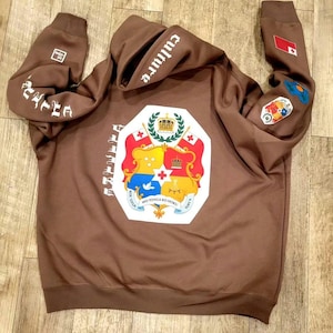 May include: Brown hooded sweatshirt with a large white crest on the front. The crest features a shield with a crown, a dove, and the words "MO TONGA KO HOKU". The words "CULTURE" and "EST. 1972" are printed on the sleeves.