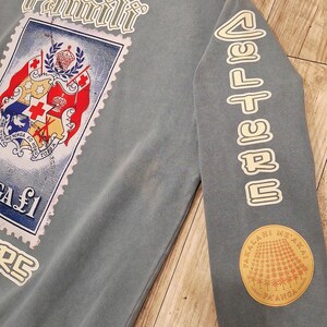 May include: A blue-grey sweatshirt with a large stamp graphic featuring a coat of arms and the word "Famili". The right sleeve has the word "CULTURE" and a circular patch with text and a sunburst design.