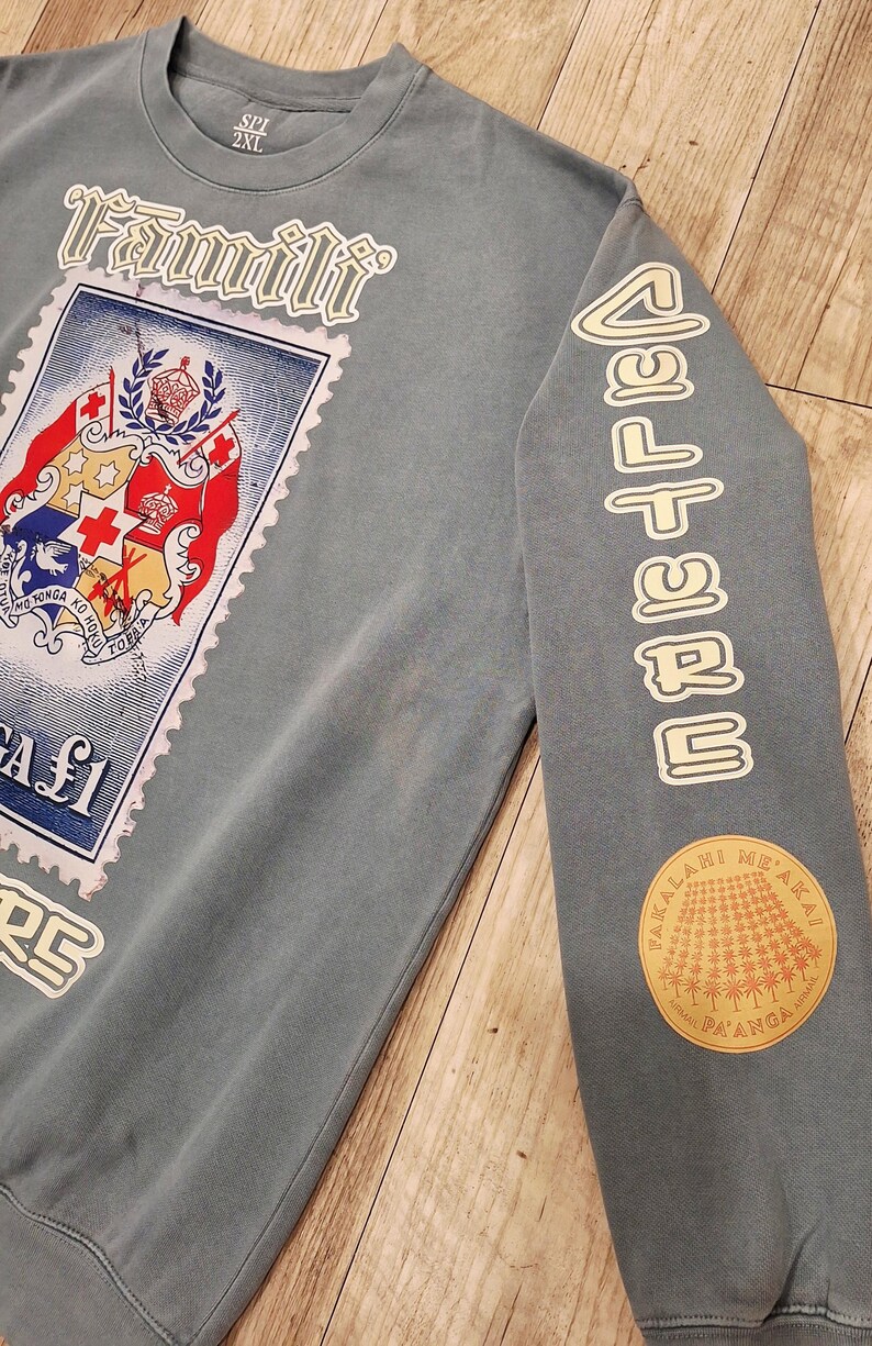 May include: A blue-grey sweatshirt with a large stamp graphic featuring a coat of arms and the word "Famili". The right sleeve has the word "CULTURE" and a circular patch with text and a sunburst design.
