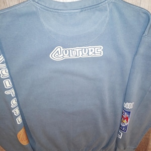 May include: A pale blue sweatshirt featuring the word "CULTURE" in white, outlined lettering on the back. The sleeves display further graphics and text. Patches with cartoon images are on the lower right sleeve.