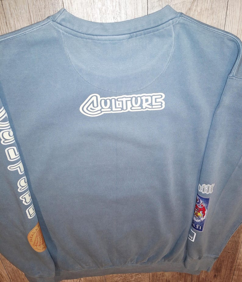 May include: A pale blue sweatshirt featuring the word "CULTURE" in white, outlined lettering on the back. The sleeves display further graphics and text. Patches with cartoon images are on the lower right sleeve.