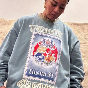 May include: A pale blue sweatshirt with a large graphic of a postage stamp featuring the Tonga coat of arms. The stamp reads "Tonga &pound;1" and "Culture" in white lettering. The word "Familie" is arched above the stamp.