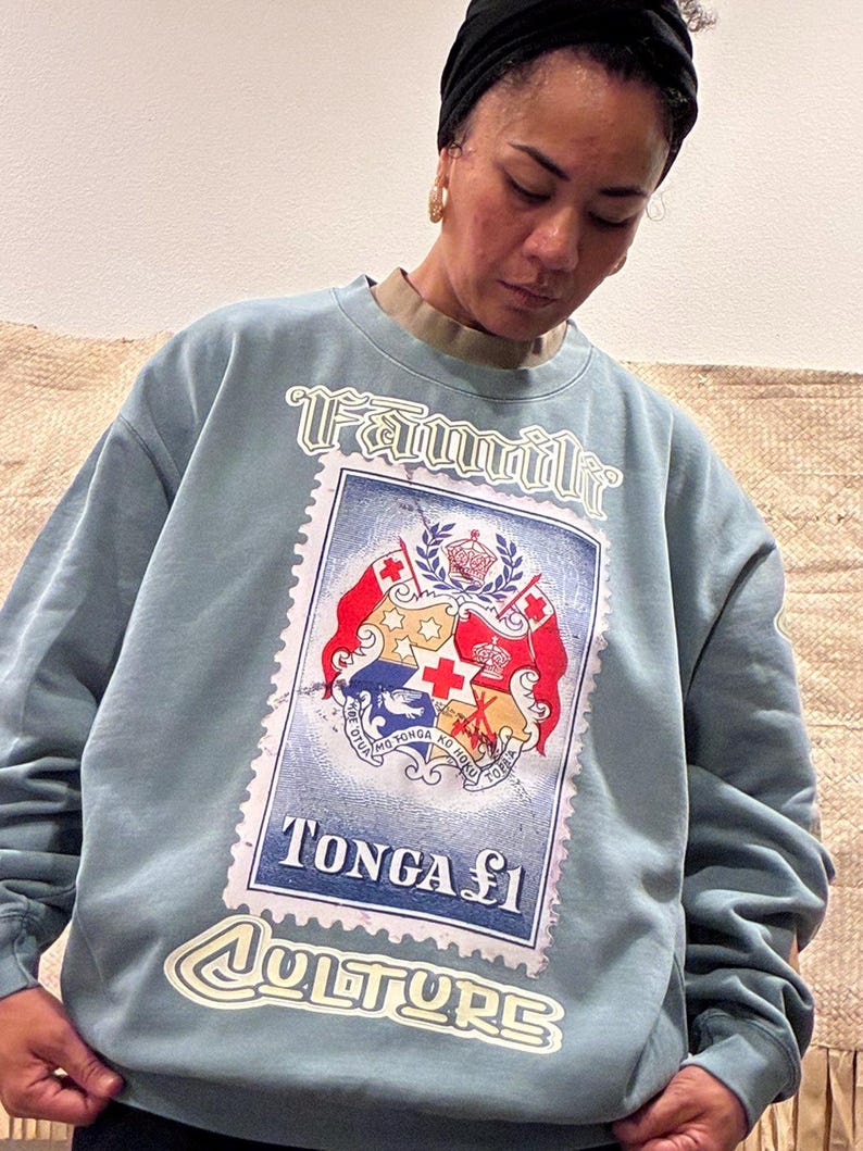 May include: A pale blue sweatshirt with a large graphic of a postage stamp featuring the Tonga coat of arms. The stamp reads "Tonga &pound;1" and "Culture" in white lettering. The word "Familie" is arched above the stamp.
