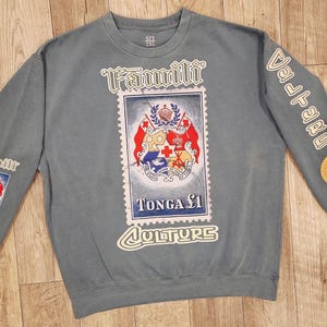 May include: A blue-grey sweatshirt with a large postage stamp design featuring the Tonga coat of arms. The words "Famili" and "Culture" are printed on the front. The sleeves have additional patches and text. The sweatshirt is size 2XL.