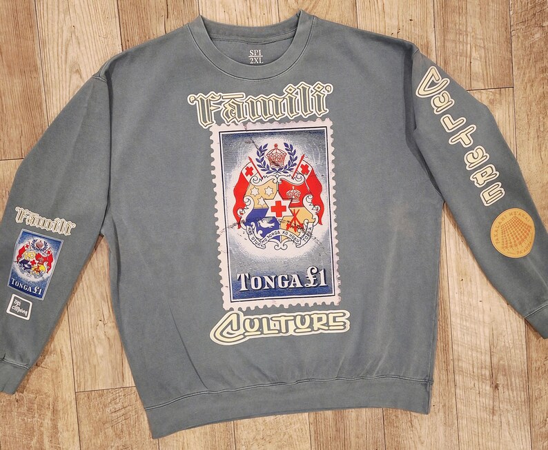 May include: A blue-grey sweatshirt with a large postage stamp design featuring the Tonga coat of arms. The words "Famili" and "Culture" are printed on the front. The sleeves have additional patches and text. The sweatshirt is size 2XL.