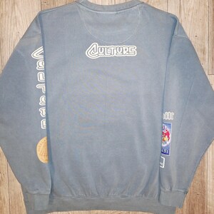 May include: A pale blue sweatshirt featuring the word "CULTURE" in white lettering on the upper back. The left sleeve has vertical white text and a circular patch. The right sleeve has a rectangular patch with a cartoon image.