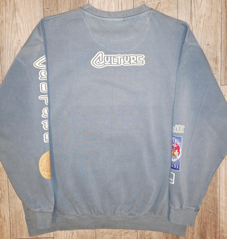 May include: A pale blue sweatshirt featuring the word "CULTURE" in white lettering on the upper back. The left sleeve has vertical white text and a circular patch. The right sleeve has a rectangular patch with a cartoon image.