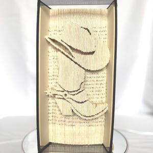 May include: A framed paper sculpture depicting a cowboy hat and boot. The artwork is crafted from book pages, with the hat and boot cut out to create a 3D effect. The frame is black.