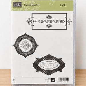 May include: A clear plastic case containing a Stampin' Up! Layered Labels stamp set. The insert displays three decorative label designs with the text CONGRATULATIONS, CREATE, and FOR YOU, along with a barcode and the number 4.