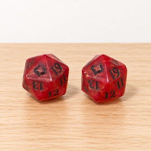 May include: Two twenty-sided dice made of translucent red resin with a marbled texture. Each die features black engraved numbers, including 11, 12, 13, and 19, with a unique symbol replacing the number 20 on each face.