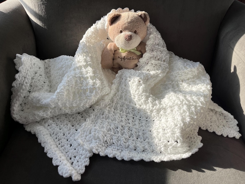 May include: White crocheted baby blanket with a scalloped edge. A brown teddy bear is sitting on top of the blanket.