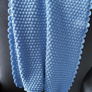 Op de afbeelding: Een lichtblauwe gehaakte deken met een textuur, verhoogd steekpatroon. De deken heeft een geschulpte rand, wat een decoratief tintje geeft. De zachte, handgemaakte deken is waarschijnlijk voor een baby of kind.