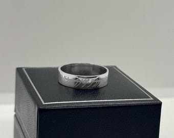 May include: A silver ring with an inscription in Elvish script. The ring is sitting on a black box.