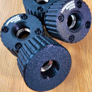 May include: Close-up of four black cylindrical objects with a textured surface and a central hole. Each object has a silver interior and is secured with black screws. The text "LEGENDARY MAKES .COM" is visible on the top of some objects.