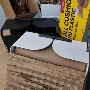 Puede incluir: Dispensadores de metal negro y blanco con un rollo de papel marrón y una caja amarilla que dice "ALL CUSHION NO PLASTIC". Los dispensadores tienen el texto "LEGENDARY MAKES" en el lateral. El papel marrón tiene un patrón de panal.