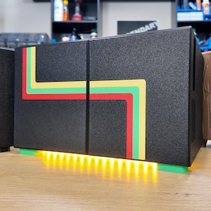 Puede incluir: Dispositivo electrónico rectangular negro con un diseño retro con rayas rojas, amarillas y verdes. El dispositivo tiene una luz amarilla brillante en la parte inferior y se asienta sobre una base verde. Sobre una superficie de madera.
