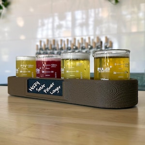 Puede incluir: Una tabla de degustación marrón con textura de madera que sostiene cuatro vasos de precipitados de vidrio de 150 ml con líquidos de colores. Una pizarra negra frontal muestra el texto escrito a mano: Hazy, Sour, Pilsner, Lager.