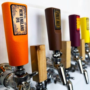 May include: A row of beer tap handles featuring various colors including orange, brown, maroon, and yellow. The orange handle is labeled New England IPA, the brown handle says Pale Ale, and the maroon handle is marked Sour. Each handle is attached to a metallic faucet mechanism.