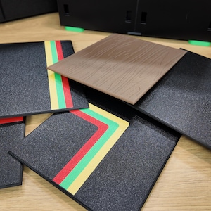 Puede incluir: Un conjunto de posavasos cuadrados negros con rayas de acento coloridas en rojo, verde y amarillo. Un posavasos tiene un patrón de vetas de madera marrón. Los posavasos están sobre una superficie de madera clara.