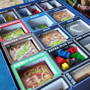 Puede incluir: Un organizador de juegos de mesa con compartimentos etiquetados. Los compartimentos contienen piezas y cartas de juego, incluyendo "MEEPLES", "RIVER", "BUILDER-TRADER" y "ABBEY-MAYOR". El organizador es azul, con acentos coloridos y componentes del juego.