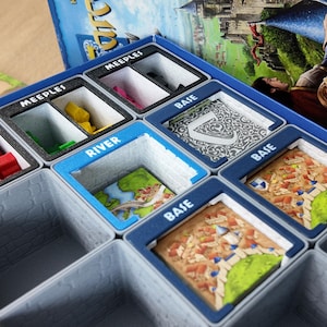 May include: A board game organizer with compartments labeled "MEEPLES", "RIVER", and "BASE". The compartments hold game pieces and tiles. The organizer is blue, gray, and black, with colorful game components visible.